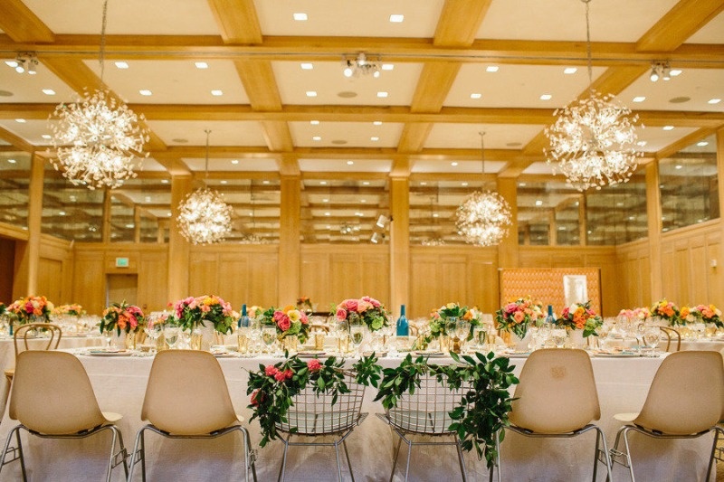 a spring wedding reception with greenery on the chairs and bright florals on the tables plus pendant lamps