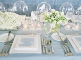 an ice blue and white winter wedding tablescape with ice blue cards and a tablecloth, white porcelain and white bloom arrangements