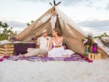 a beach wedding picnic with a teepee, colorful blooms, bright glass lanterns, a colorful blanket and pallets as stands