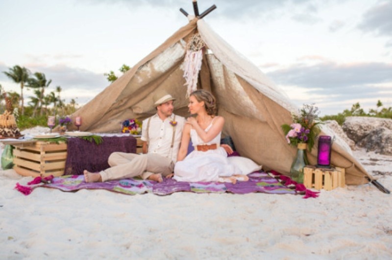 A beach wedding picnic with a teepee, colorful blooms, bright glass lanterns, a colorful blanket and pallets as stands