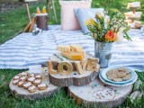 a pastel wedding picnic with a striped blanket, layered pillows, bright blooms, lots of delicious sweets served on wood slices