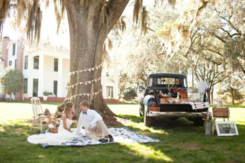 A vintage rustic wedding picnic with a paper bunting, some photos on the tree, a blanket and a vintage car as a sweets table