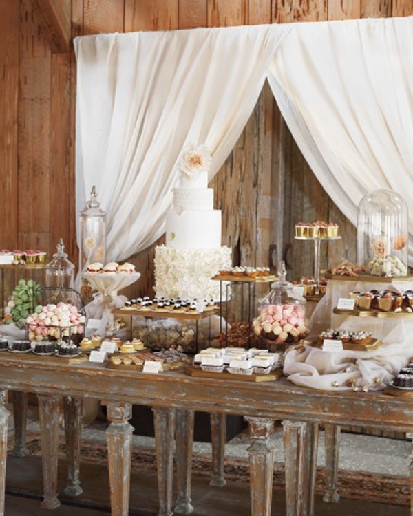 A rustic food display of a shabby chic table, vintage stands and cloches with sweets and some curtains behind the table