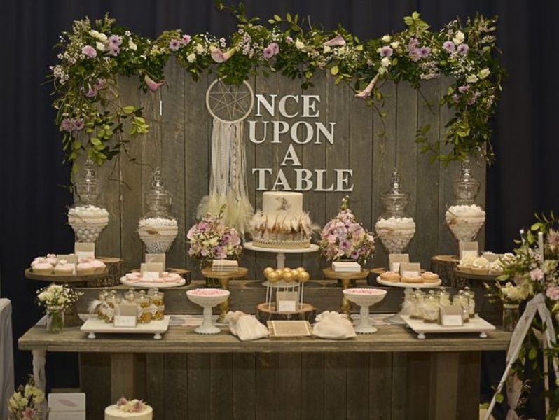 A refined rustic dessert table of a wooden table and a wooden screen with greenery and a dream catcher, some pastel and neutral blooms and stands and glass jars with sweets