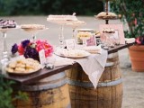 a vineyard dessert table with two barrels and a tabletop, bright blooms and glass and wooden sweets stands