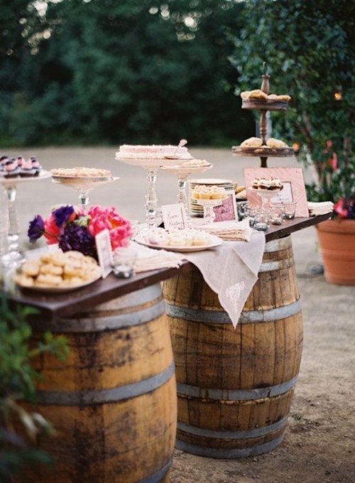 a vineyard dessert table with two barrels and a tabletop, bright blooms and glass and wooden sweets stands