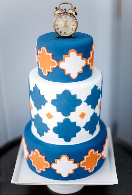A bright Moroccan style wedding cake in blue, orange and white, with a Moroccan pattern and a clock on top is a cool and lovely idea to try