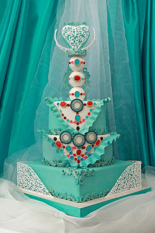 A whimsical turquoise wedding cake with three tiers and three balls on top, with a heart and some lace detailing plus some ruffles and bold spots