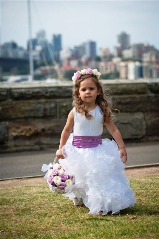 A flower girl wearing a sleeveless white maxi dress with a radiant orchid sash and a matching floral crown plus a basket with blooms