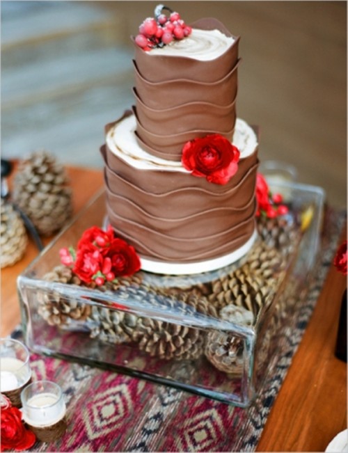 a whimsical chocolate ruffle wedding cake with red blooms and berries placed on a clear stand with pinecones is a lovely and bold idea to rock