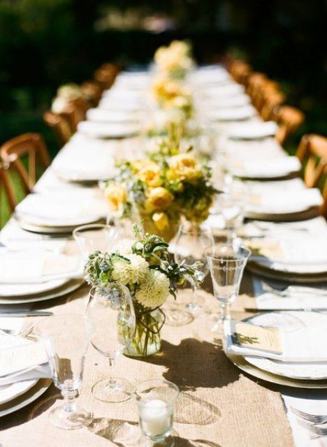 a simple rustic table setting with a white tablecloth, a burlap runner, yellow blooms in vases