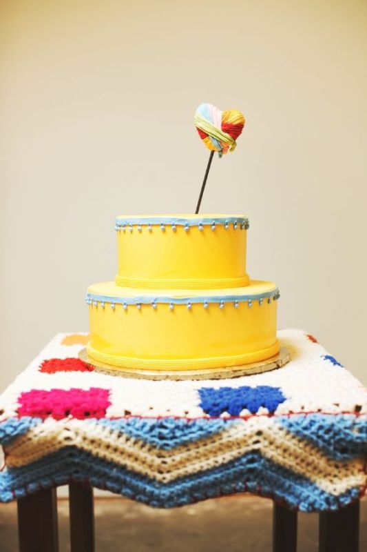 A bold and cool yellow wedding cake with blue ribbons with bells and a bold yarn heart topper is a whimsy dessert idea
