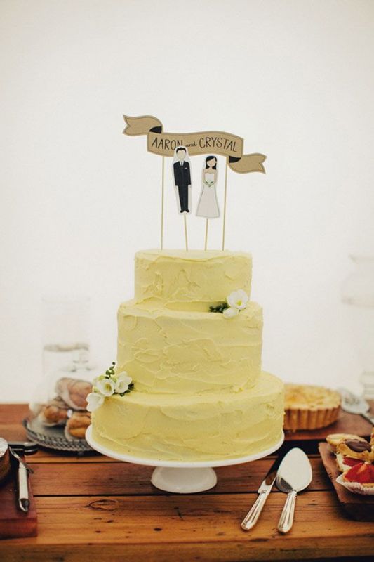A pale yellow textural wedding cake with white blooms and cake toppers, with the couple's names is a cool idea