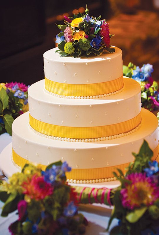 A white patterned wedding cake with yellow ribbons, beads and bold blooms on top is a bright and fun idea
