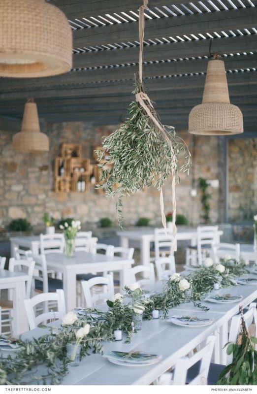 A olive greenery chandelier and matching table runners with candles and white blooms is a cool rustic setup for a wedding venue