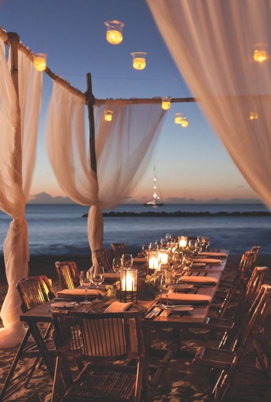 A beach wedding reception space with suspended and usual candle lanterns and breezing curtains plus a gorgeous sea view