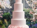 a large light pink wedding cake with sleek tiers and a ruffle one, with pink sugar blooms on top is a very chic and beautiful idea