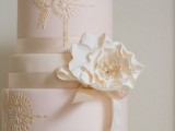 a light pink and white wedding cake with multiple tiers, with gold patterns, a white sugar bloom is a lovely idea for a sophisticated wedding