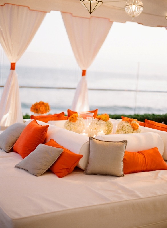 a neutral wedding lounge with curtains, neutral and bright orange pillows that add color and interest to the space
