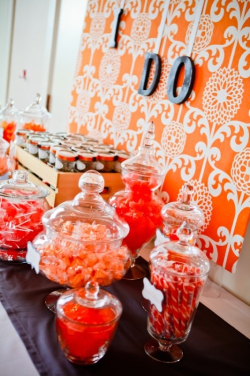 a bold orange wedding candy table with lots of candies in jars, an orange printed wallpaper wall is an amazing solution