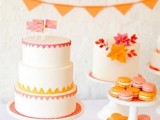 a cool wedding sweets table with an orange banner, white wedding cakes with orange and red detailing, orange and pink macarons