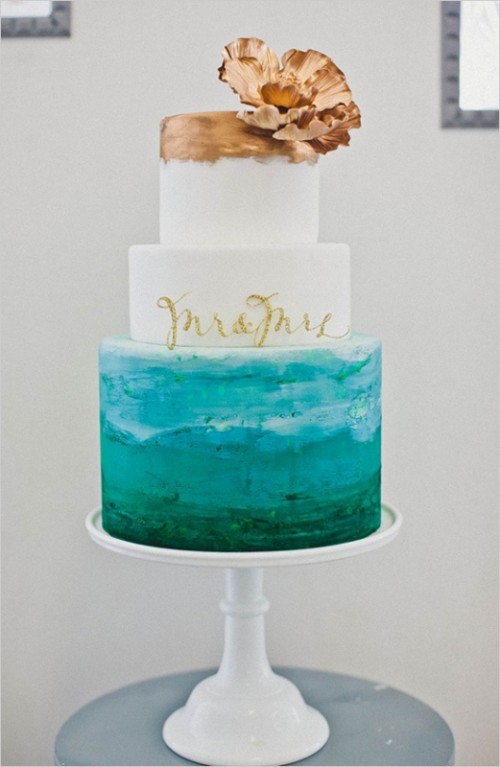 an ocean-inspired wedding cake with white, white and gold and turquoise tiers, the latter showing the sea, a gold sugar bloom on top