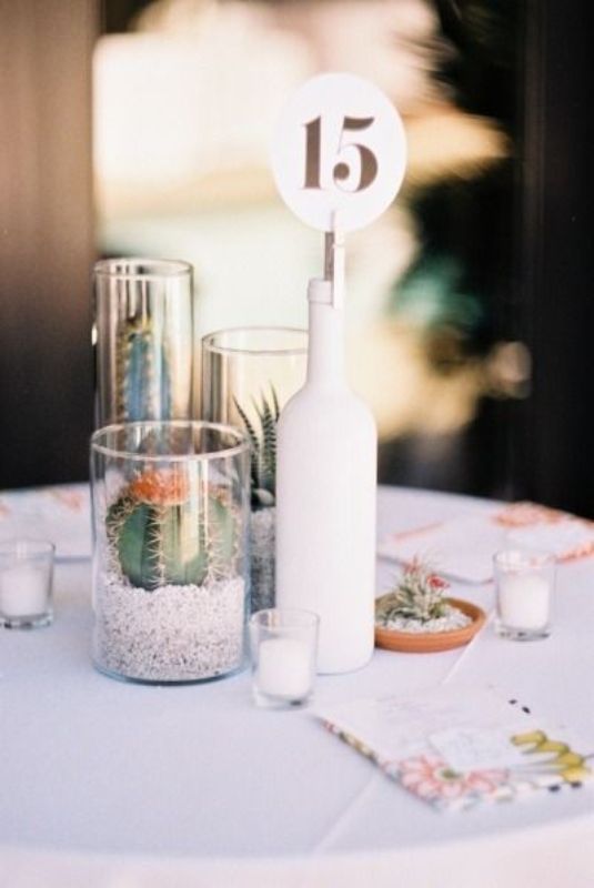 A modern cluster wedding centerpiece of a white bottle with a table number, glasses with cacti and succulents and mini candles around