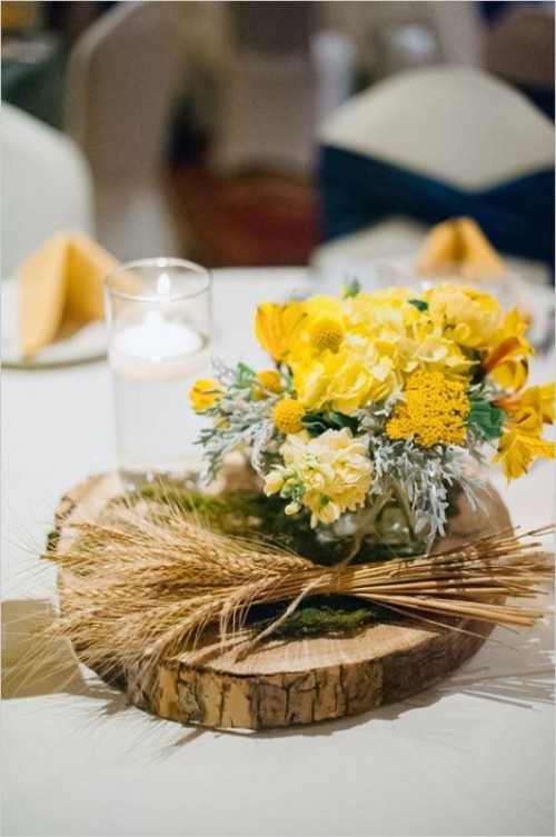 a bright rustic wedding centerpiece of a wood slice, wheat, moss and yellow blooms and pale greenery