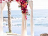 a colorful tropical wedding arch done with neutral fabric and bright blooms and greenery and a gorgeous ocean view