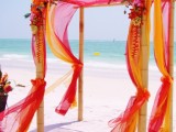 a super colorful tropical wedding arch decorated with yellow, orange and red fabric and matching bright blooms is wow