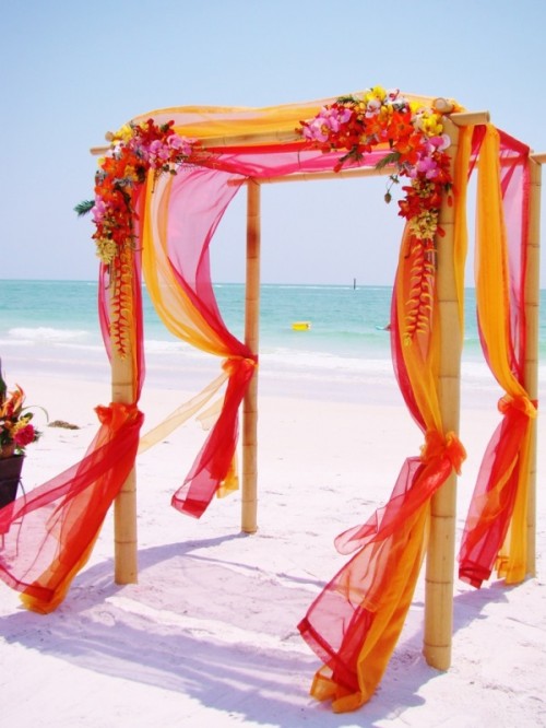 a super colorful tropical wedding arch decorated with yellow, orange and red fabric and matching bright blooms is wow