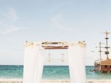 a refined tropical wedding arch covered with white fabric, neutral blooms and hanging lanterns