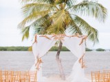 a chic tropical wedding ceremony space with an arch with neutral fabric and driftwood and a palm tree in the backdrop