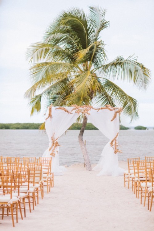 a chic tropical wedding ceremony space with an arch with neutral fabric and driftwood and a palm tree in the backdrop