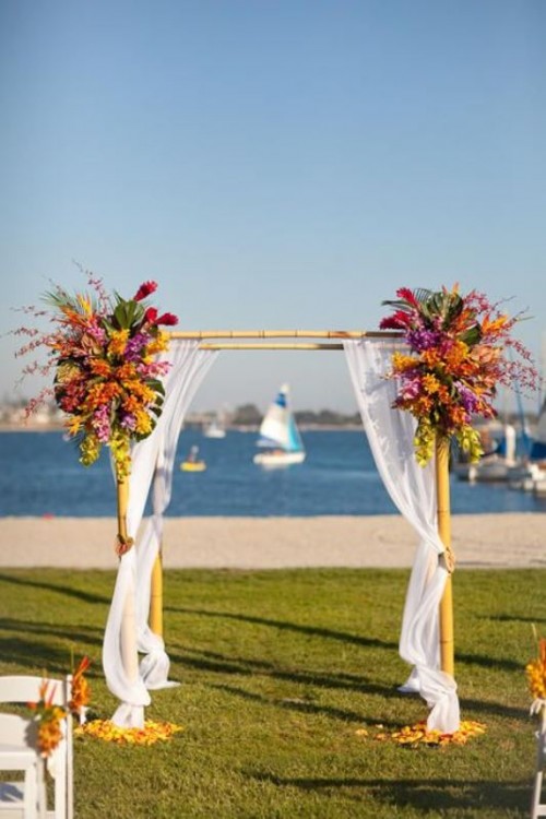 a bamboo wedding arch decorated with white curtains and with super bright blooms and greenery