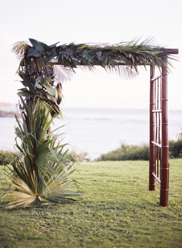 A dark bamboo wedding arch covered with all kinds of tropical leaves on one side is bold and cool