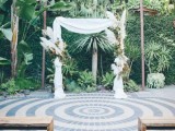 a tropical wedding arch decorated with white fabric, pampas grass and leaves in brown and green