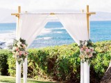 an elegant tropical wedding arch decorated with white fabric, greenery, neutral and pastel flowers