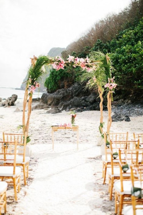 a driftwood wedding arch decorated with palm leaves and pink flowers with an ocean view is awesome