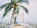 a breezy tropical wedding ceremony space with an arch covered with white fabric, bright petals on the ground and a large palm tree in the backdrop