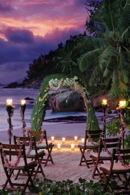 a natural tropical wedding ceremony space done with an arch covered with palm leaves, lots of candles and floral garlands on the chairs