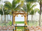 a refined tropical wedding ceremony space with a yellow arch covered with fabric and blooms, accented chairs and petal patterns on the ground