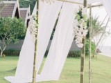 a bamboo tropical wedding arch decorated with white blooms and airy white fabric is very breezy and cool