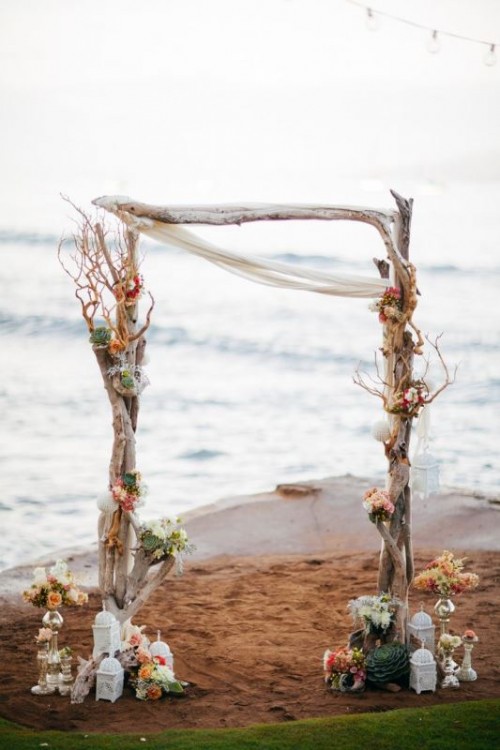 a driftwood wedding arch decorated with neutral blooms, pale greenery and succulents, candle lanterns and large succulents around