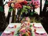 a colorful tropical wedding tablescape with a green tablecloth and pink placemats, a bright floral and fruit table runner and a bold installation overhead