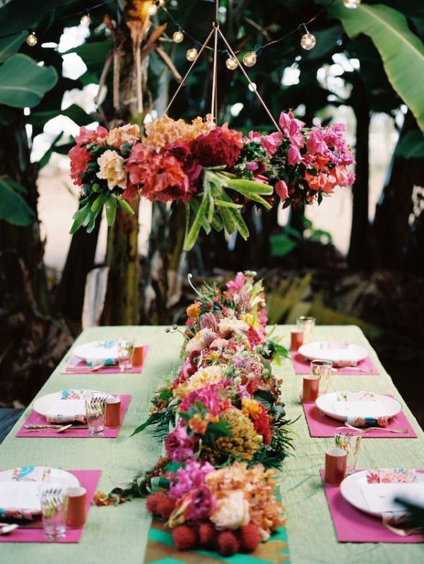 A colorful tropical wedding tablescape with a green tablecloth and pink placemats, a bright floral and fruit table runner and a bold installation overhead