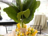 a bold tropical wedding tablescape with mustard blooms and oversized tropical leaves, woven chargers, candles, green napkins