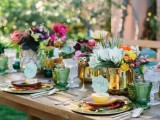 a colorful wedding tablescape with gold chargers, cutlery and vases, green glasses and bright blooms