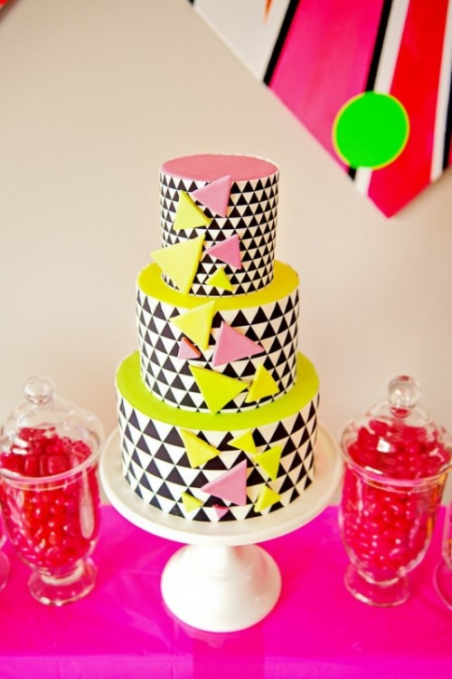 a round wedding cake with black and white triangles covering the tiers and bold pink and neon yellow tops and triangles all over the cake