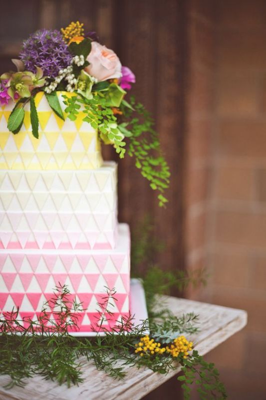 a colorful modern wedding cake with an ombre effect, with yellow, white and pink triangle tiers and super bold blooms and greenery on top the cake